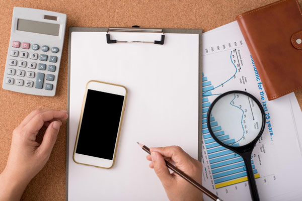 Top view of a workspace with a clipboard, smartphone, calculator, financial charts, magnifying glass, and a person's hands holding a pen.